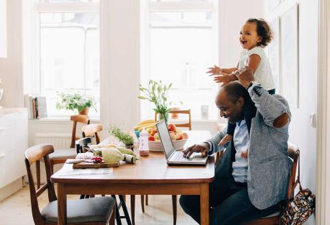 A man sitting at a desk in his living room with his toddler daughter laughing on his shoulders.